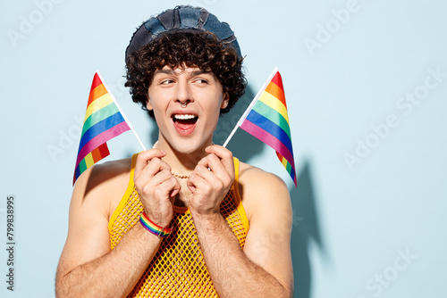 Young surprised happy gay Latin man wear mesh tank top hat clothes hold in hand two striped flags look aside isolated on plain blue background studio portrait. Pride day June month love LGBT concept.
