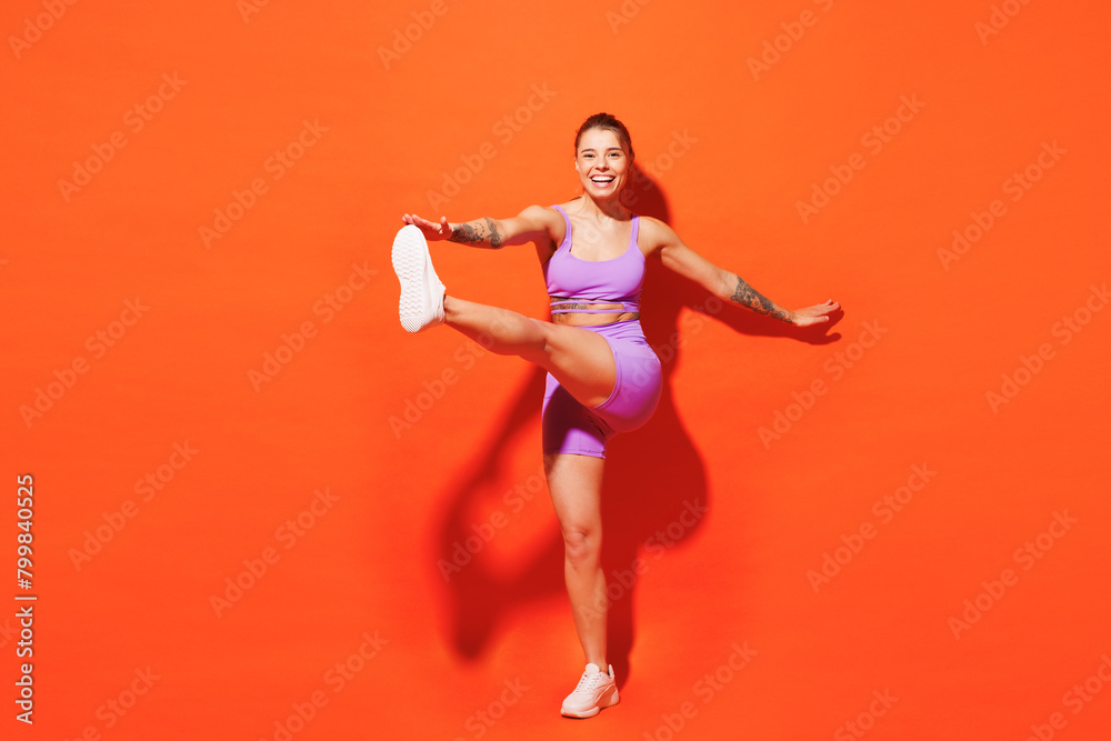Full body young fun fitness trainer woman sportsman wear purple top clothes train in home gym raise up leg do stretch hand exercise isolated on plain orange background. Workout sport fit abs concept.