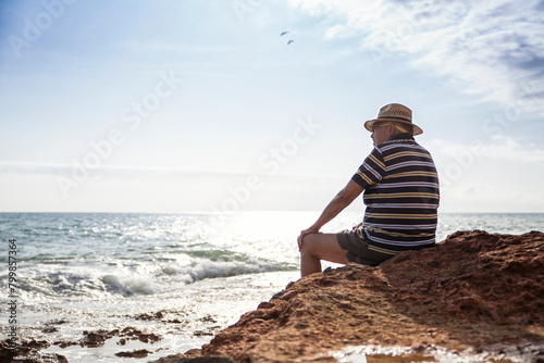 Italy, Sicily, Cava D'aliga, senior man sitting at the coast and looking to the sea photo