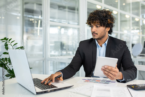 A focused young man multitasks with a laptop and tablet at a desk surrounded by light-filled modern office space. He exudes professionalism and efficiency in a corporate environment. photo