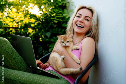 Happy woman with kitten and laptop sitting by plants in balcony