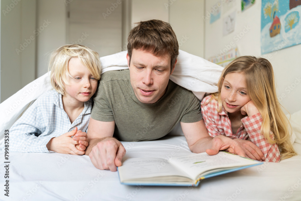 Father reading book to son and daughter lying on bed at home Father reading book to son and daughter lying on bed at home