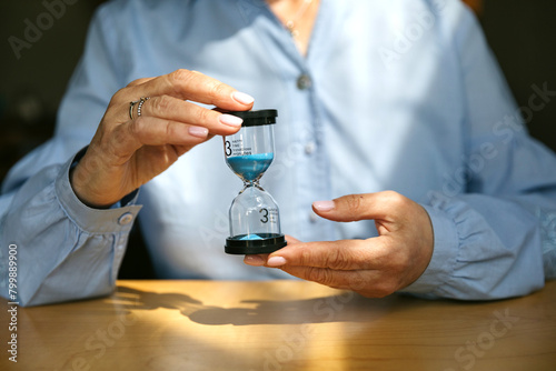 Unrecognizable female hands holding transparent hourglass with blue sand on blue background. Conceptual photo about time, business and time management.