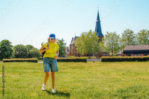 A woman in a yellow shirt and blue shorts walks through a grassy field against the backdrop of an ancient European castle