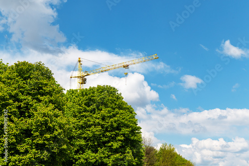 Industrial construction crane behind the trees against the sky with white clouds.