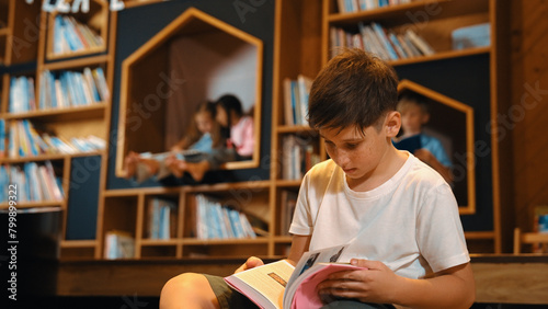 Fotografie Caucasian boy reading a book while group of smart students sitting at library