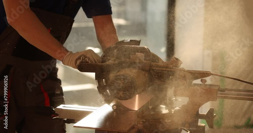Worker uses sawing machine to cut wooden board at workplace in semi-lit room. Male specialist prepares material for parquet laying in renovated room slow motion