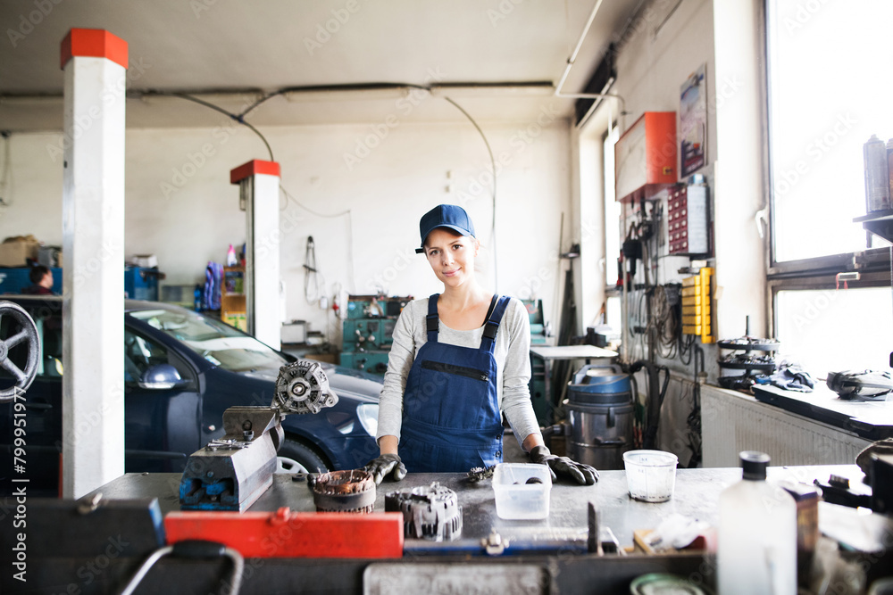 Female auto mechanic repairing, maintaining car. Beautiful woman standing in a garage, wearing blue coveralls.