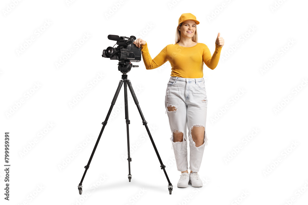 Full length portrait of a female camera operator with a camera on a tripod stand gesturing thumbs up