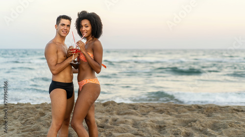 Multiethnic couple posing on the beach holding cocktails - Romantic seaside ambiance - Celebrating togetherness with ocean waves in the background - copy space.