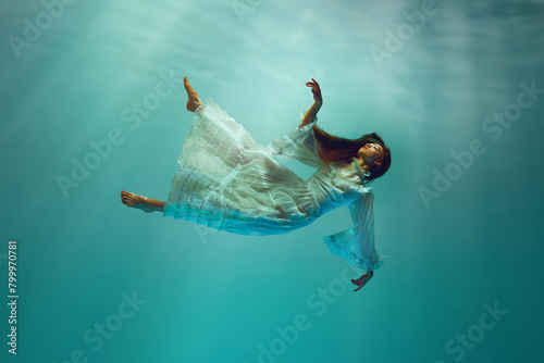 Finding inner peace. Serene underwater moment with elegant young girl in tender white dress levitating with calm expression underwater. Concept of surrealism, beauty, mystery and fantasy, freedom photo