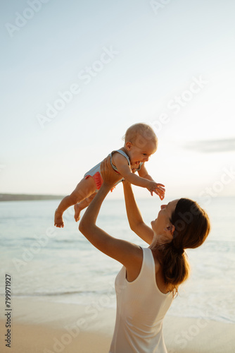 Young mother throws up baby in the sky in front of the sea, summer outdoors. Happy mom and cute smiling baby girl. Positive human emotions, feelings, natural lifestyles. Family background