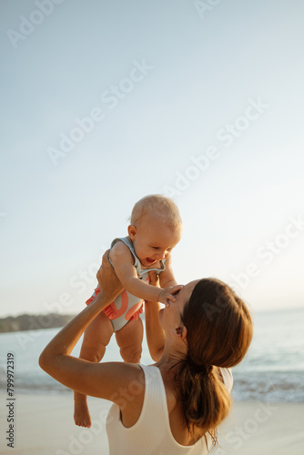 Young mother throws up baby in the sky in front of the sea, summer outdoors. Happy mom and cute smiling baby girl. Positive human emotions, feelings, natural lifestyles. Family background