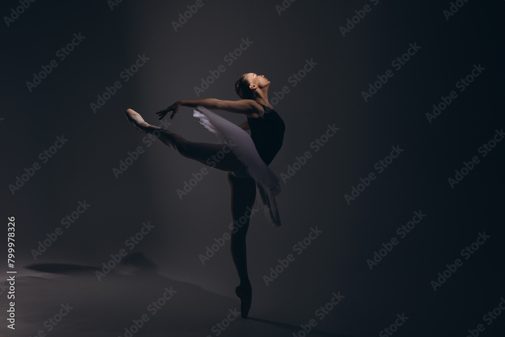 Obraz premium Young ballerina in elegance white tutu and pointe shoes performing Arabesque pose against dark background.