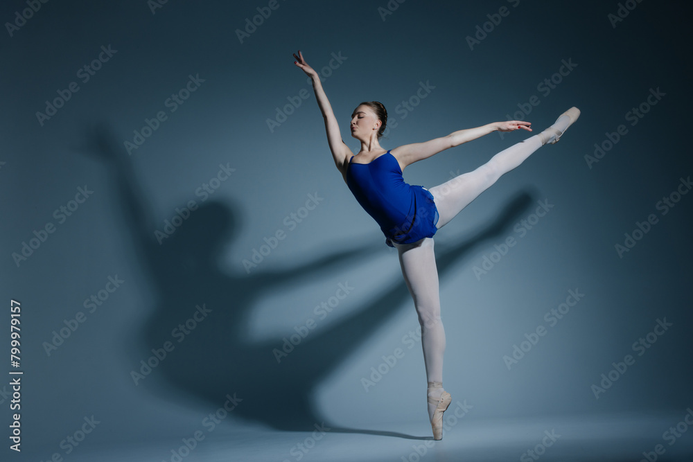 Young ballerina in bodysuit and pointe shoes performing Arabesque pose against of herself shadow on blue background.