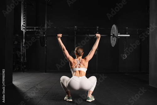 Rear view of woman performing overhead squat pressing barbell up, standing up. Routine workout for physical and mental health.