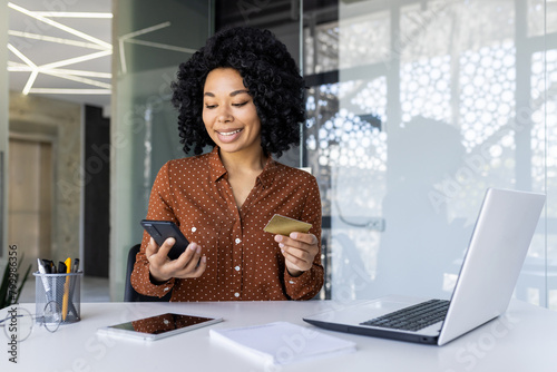 Confident African American businesswoman using a smartphone and credit card for online transactions at her modern office workspace.