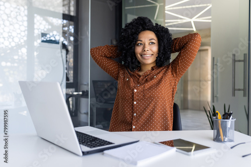 A young African American businesswoman with natural curly hair, dressed in a stylish polka dot blouse, relaxes at her workspace filled with light, exuding confidence and comfort.