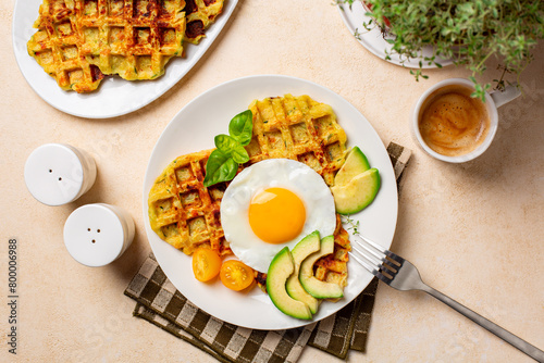 Homemade breakfast or brunch. Fried egg with belgian waffles made with cheese and vegetables, zucchini, onion, carrot, served with avocado and yellow tomato. Cup of coffee drink.