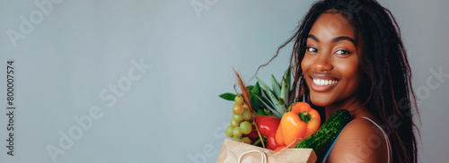 Portrait of a young African American woman - a happy girl with long hair and a snow-white smile holding a paper bag of vegetables on a gray banner background.