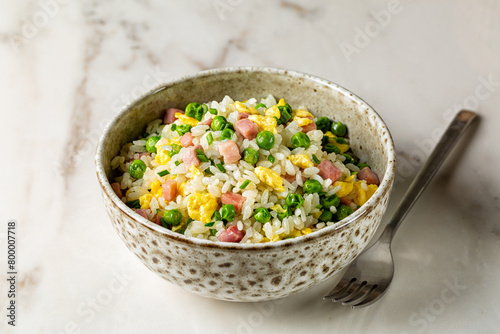 Cantonese Fried Rice and egg with green pea, ham steak, spring onion, ribe long rice in a beige bowl on a white marble table, served with fork. Eurasian cuisine.