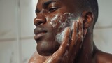 A man with a soapy face, gently washing his skin in a shower, with a focused and serene expression.