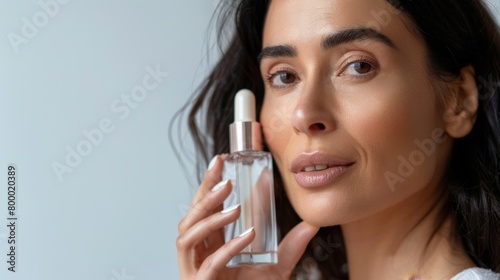 Woman with long dark hair holding a clear bottle of skincare product applying it to her face with a gentle touch against a soft-focus background. photo
