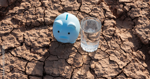 Heat-cracked clay in the desert, a glass of drinking water and a piggy bank