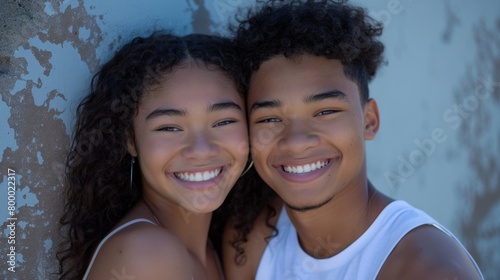 Two young individuals possibly siblings smiling brightly at the camera their joy radiating against a backdrop of a weathered blue wall.