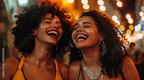 Two women with curly hair laughing joyously at night with blurred lights in the background.