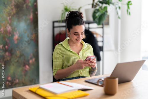 A content Hispanic woman in a green-striped shirt enjoys her smartphone at a modern workspace, reflecting a seamless blend of technology and lifestyle photo