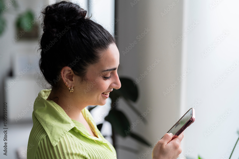 A smiling woman engaged in a pleasant phone conversation near a bright window, adorned in a lime-green shirt. A smiling woman engaged in a pleasant phone conversation near a bright window, adorned in a lime-green shirt.