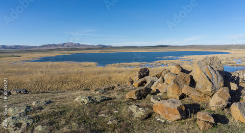 Huge stones and lake with clear water. Mountains and agricultural fields. Bright blue sky