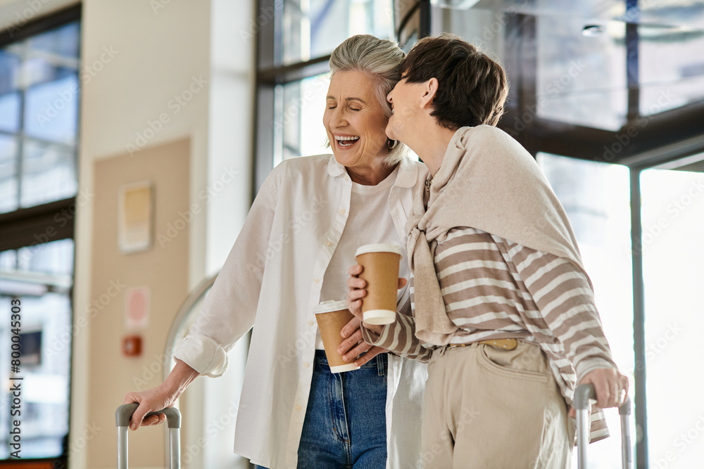 Obraz premium Two senior lesbian women holding luggage and sipping coffee.
