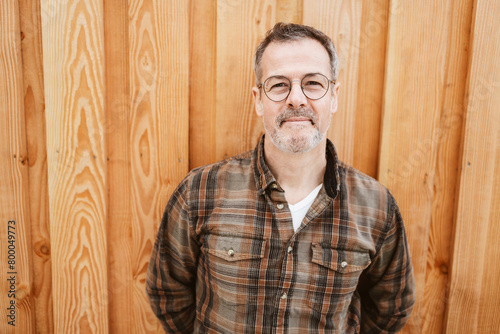 Mature man with glasses standing outdoors in front of a wooden wall, smiling confidently