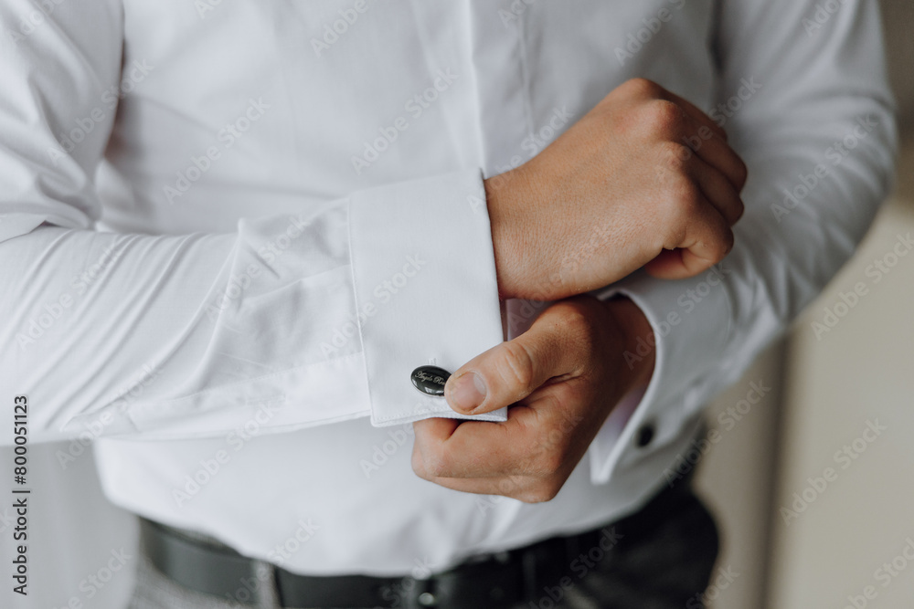 A man is getting dressed in a white shirt and black pants. He is adjusting his cuff and holding a button in his hand