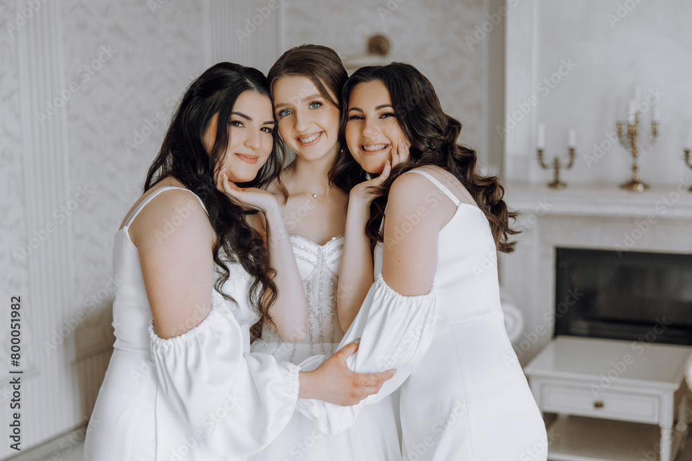 Three women in white dresses are posing for a picture. They are all smiling and seem to be happy. The room they are in has a fireplace and a few candles, giving it a cozy and warm atmosphere