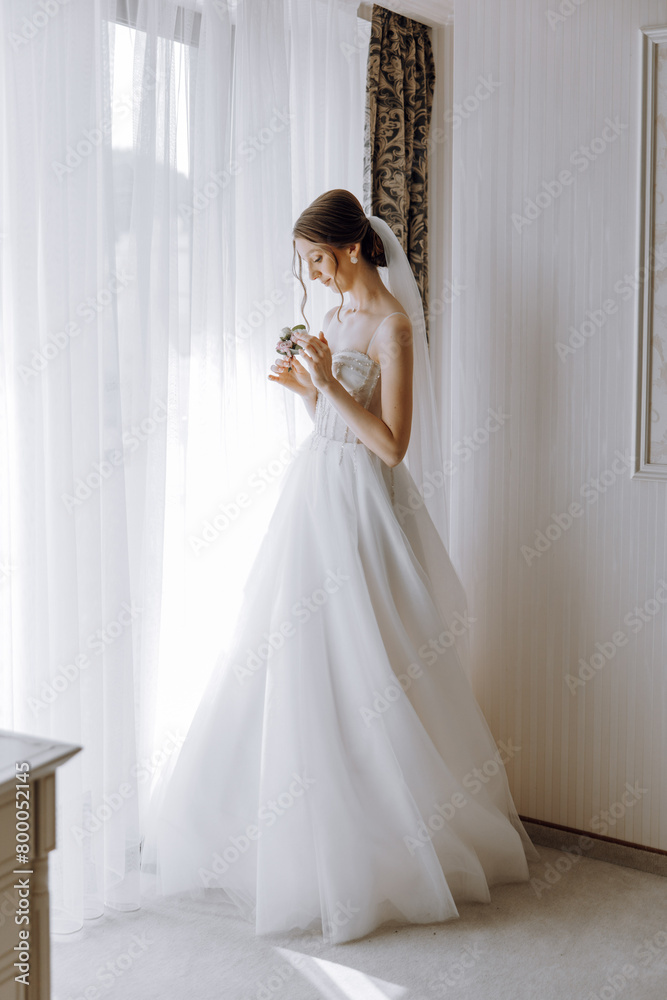 Fototapeta premium A bride is standing in front of a window, holding a camera and looking out. She is wearing a white dress and a veil