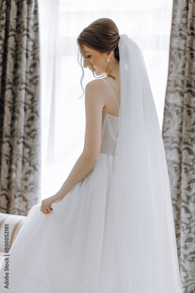 A woman in a white dress is standing in front of a window. She is wearing a white veil and earrings
