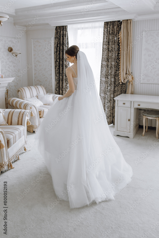 A woman in a white wedding dress is standing in front of a desk. The room is decorated with a lot of furniture, including two chairs and a desk. The woman is wearing a veil
