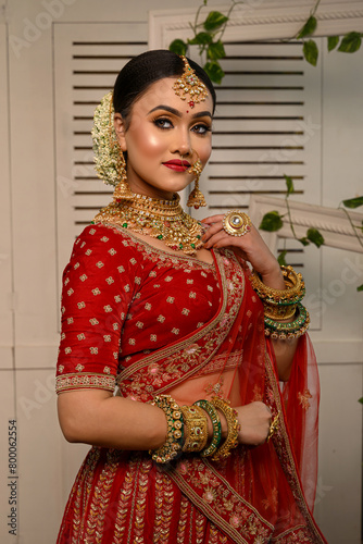 Stunning Indian bride adorned in a traditional red bridal lehenga gracefully wears heavy gold jewellery and a veil and smiles tenderly in studio lighting. Wedding fashion and lifestyle.