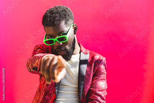 Young non-binary person wearing sunglasses and pointing against pink background photo