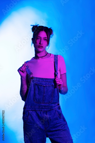Beautiful young woman in bib overalls standing against illuminated background