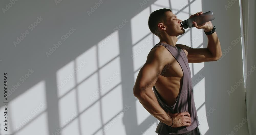 A fit 23-year-old man hydrates with a water bottle after a workout session in a brightly lit studio, showcasing an athletic physique. Athletic Man Drinking Water After Training in Studio