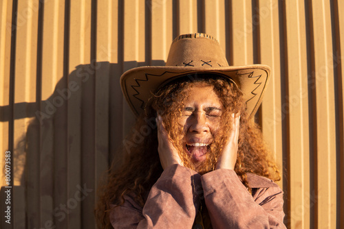 Curly haired woman wearing cowboy hat shouting in front of orange metal wall photo