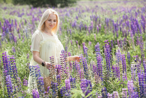 A cute pregnant girl in light underwear and a peignoir, with blond short hair, walks and rests in a picturesque meadow with purple blossoms.