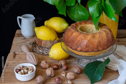 Aerial view of lemon bundt on wooden table with lemons, nuts and green leaves, black background, horizontal