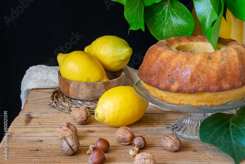 Closeup of lemon bundt on wooden table with lemons, nuts and green leaves, black background, horizontal, with copy space