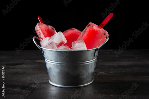 View of metal bucket with watermelon and ice lollies, on table and dark background, horizontal, with copy space