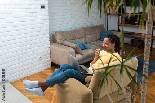 Cheerful young black woman with dreadlocks watching comedy movie on laptop at home, sitting on chair by coffee table, eating popcorn and laughing. African american lady having fun during isolation © Srdjan